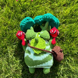 Könnte beinhalten: Handgefertigte Häkelpuppe in Grün und Weiß, die eine Figur mit baumartigem Kopf und roten Akzenten darstellt. Sie hat einen kleinen braunen Rucksack und liegt auf grünem Gras.