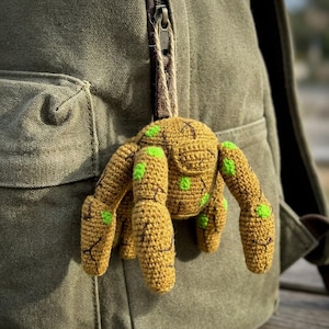 May include: A handmade crochet spider in shades of mustard yellow and green, hanging from a string. The spider has a round body and eight legs, with green spots. The spider is attached to a green backpack.