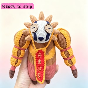 May include: A crochet plush toy featuring a white face with dark blue markings, two brown button eyes, and a spiky tan crown. The body is brown and tan with large yellow arms and a red chest panel featuring gold embroidered symbols and Ready to ship text.