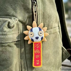 Puede incluir: Un llavero hecho a mano con forma de sol de ganchillo con una cara sonriente, detalles azules y blancos y una etiqueta roja con caracteres chinos. El llavero está sujeto a una cremallera de mochila verde.