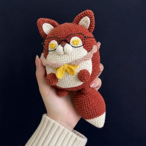 Op de afbeelding: Een rond, gehaakt vossenknuffeltje van roestkleurig garen met een crèmekleurige buik en een staart met een witte punt. De vos heeft gele ogen, een kleine bruine neus, een lichtroze sjaal met een gele strik en een dunne zwarte bril.