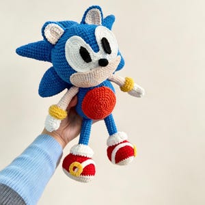 May include: A blue, white, red, and yellow crocheted plush toy shaped like a cartoon hedgehog. The figure features large white eyes with black pupils, a small black nose, a round orange belly, and red shoes with white accents and yellow rings.