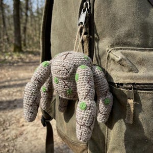 May include: A crocheted spider charm in shades of beige and green, suspended from a backpack. The spider features eight legs and green detailing. The charm is attached with a natural fibre string, highlighting a handmade craft.