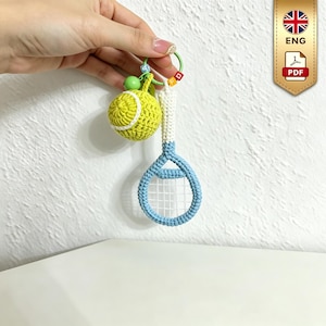 Puede incluir: Un llavero de ganchillo que presenta una pelota de tenis amarilla y una raqueta de tenis azul claro con mango blanco. Los artículos están unidos a una anilla verde decorada con pequeñas cuentas de letras coloridas, creando un accesorio detallado.