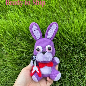 May include: A handmade purple crochet bunny plushie with a red bow tie and a small guitar. The bunny has large, round eyes and is set against a backdrop of green grass.
