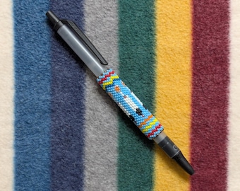 May include: A gray pen with a black tip and clip. The pen's barrel is adorned with a beaded design in blue, white, orange, yellow, and red. The pen rests on a striped background of various colors.