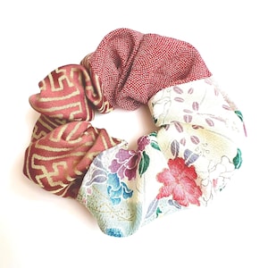 Może przedstawiać: Kolorowa gumka do włosów z różnych tkanin. Posiada sekcje z czerwonej tkaniny ze złotymi geometrycznymi wzorami, białymi kropkami i kwiatowymi wzorami w kolorze czerwonym, fioletowym i niebieskim na kremowym tle.