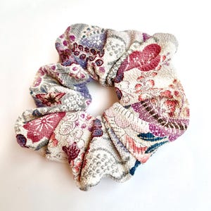 May include: A floral scrunchie in shades of purple, pink, and gray. The fabric features a detailed pattern of flowers and other shapes. The scrunchie is made of a soft, textured material.
