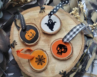 May include: Four Halloween-themed wooden ornaments with black and orange designs. Each ornament is a small, round slice of wood with a different spooky image: a pumpkin, a ghost, a skull, and a spider web. They have black and white or orange ribbons.