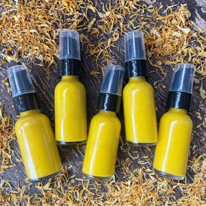 May include: Six amber glass bottles with gold lids filled with a yellow liquid. The bottles are arranged in a semi-circle on a surface covered in dried yellow flower petals.