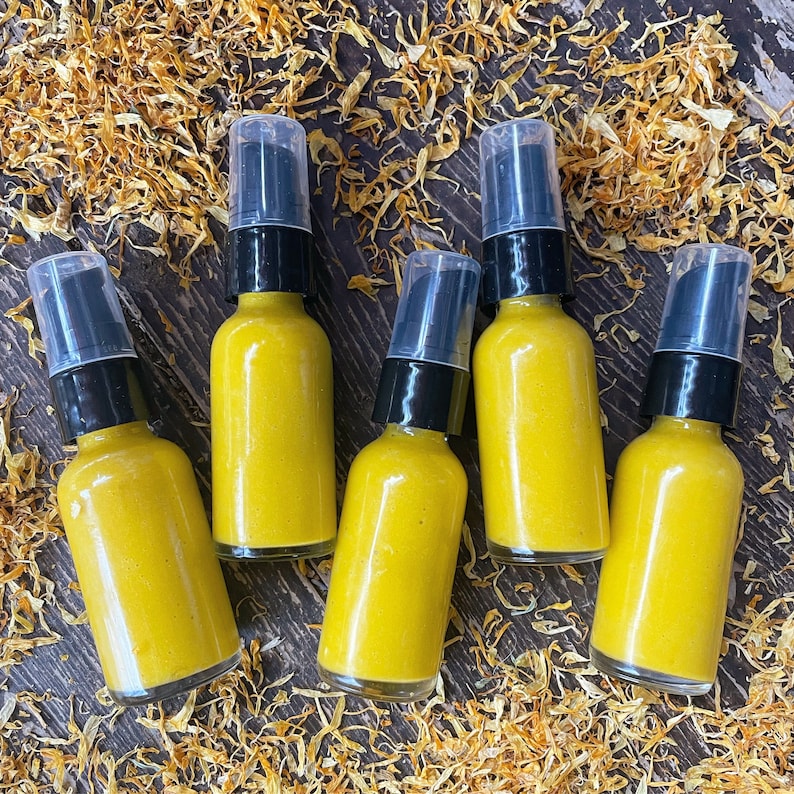 May include: Six amber glass bottles with gold lids filled with a yellow liquid. The bottles are arranged in a semi-circle on a surface covered in dried yellow flower petals.