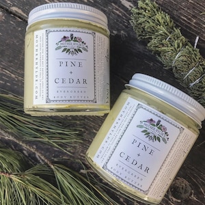 May include: Two glass jars of body butter with white lids. The jars are labelled "Mother Hyldes" and "Pine + Cedar Evergreen Body Butter".