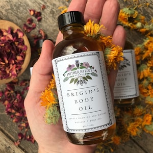 以下が含まれることがあります： 手で持たれた、黒いキャップとテキストと花のイラストが描かれた白いラベルが付いた、Brigid's Body Oilの透明なガラス瓶。「Mother Hylde's Herbal」と「Gently Warming and Moving Massage + Body Oil」と書かれています。ボトルには120mlが入っています。