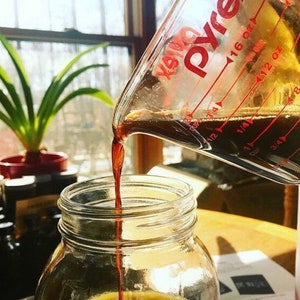 May include: A clear Pyrex measuring cup pouring a dark liquid into a clear glass jar. The measuring cup has red markings for ounces and cups. A green plant in a red pot is in the background.