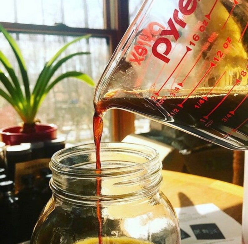 May include: A clear Pyrex measuring cup pouring a dark liquid into a clear glass jar. The measuring cup has red markings for ounces and cups. A green plant in a red pot is in the background.