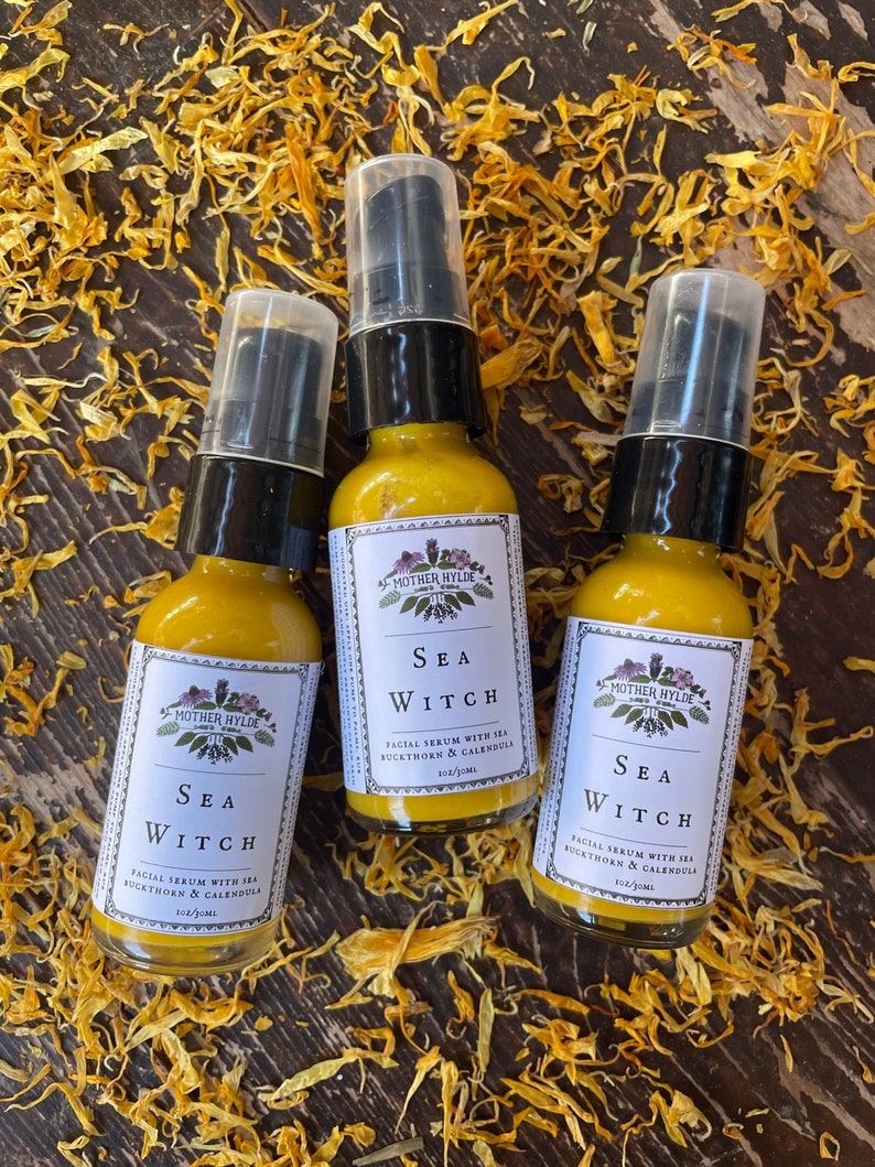 May include: Three amber glass bottles with black caps and clear pump tops. Each bottle is labeled "Sea Witch" and contains a facial serum with sea buckthorn and calendula. The bottles are sitting on a bed of dried yellow flower petals.