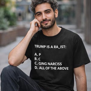 May include: A black short-sleeved t-shirt featuring white text that reads: TRUMP IS A RA_IST: A. P, B. C, C. GING NARCISS, D. ALL OF THE ABOVE. The shirt has a crew neckline and a standard fit, displaying a multiple-choice question graphic on the front.