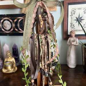 May include: A decorative wall hanging featuring a central wooden post with a carved rune, adorned with a large olive green burlap bow, a woven twig wreath, brown satin ribbons, twine, artificial green vines, and two small metal bells.