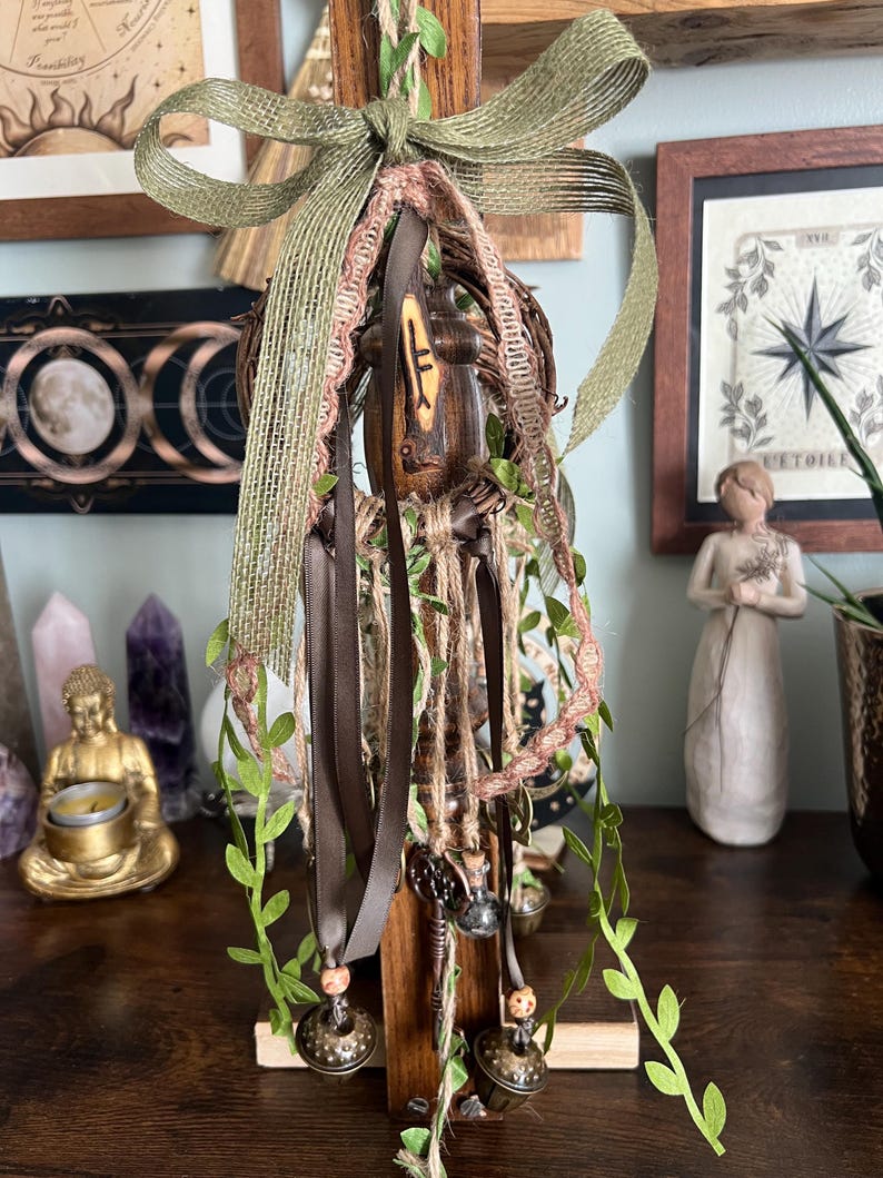 May include: A decorative wall hanging featuring a central wooden post with a carved rune, adorned with a large olive green burlap bow, a woven twig wreath, brown satin ribbons, twine, artificial green vines, and two small metal bells.