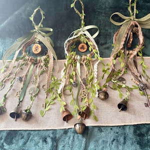May include: Three hanging wall decorations featuring circular woven vine frames adorned with green faux ivy, jute twine, and olive green ribbons. Each piece includes dangling metal charms, keys, and bells, with small wooden discs displaying symbols.
