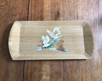 May include: A wooden serving tray with a rectangular shape and rounded edges. The tray features a painted design of three white swans flying over a body of water with reeds.