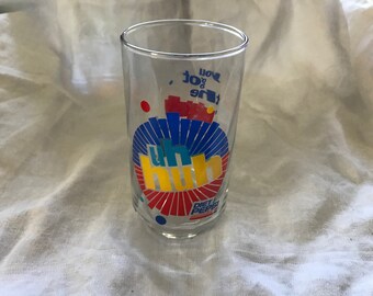 Può includere: Un bicchiere di vetro trasparente con un logo "uh huh" blu, giallo e rosso e il testo "the you got it" stampato sul lato. Il bicchiere ha anche il logo Diet Pepsi stampato sul fondo.