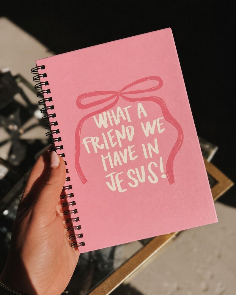 May include: A pink spiral-bound notebook featuring the text "WHAT A FRIEND WE HAVE IN JESUS!" in white script, with a pink bow design. The notebook is held in a hand.