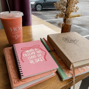 May include: A stack of journals and a Bible are on a wooden table, with a pink notebook on top that reads "WHAT A FRIEND WE HAVE IN JESUS!". A smoothie in a clear cup with a straw and a vase of dried flowers are also present. The scene is near a window.