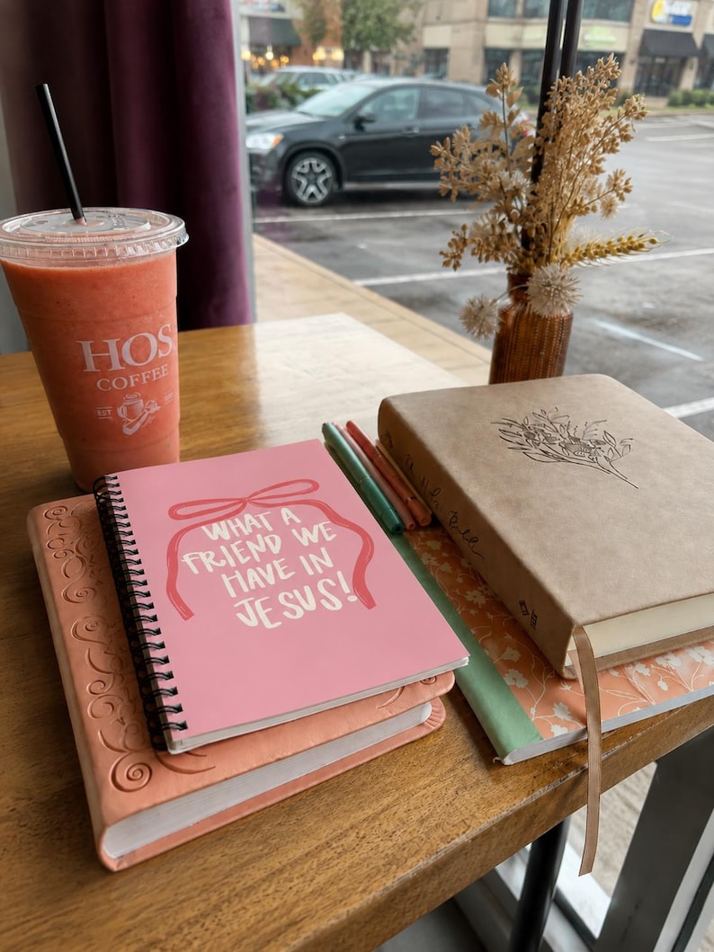 May include: A stack of journals and a Bible are on a wooden table, with a pink notebook on top that reads "WHAT A FRIEND WE HAVE IN JESUS!". A smoothie in a clear cup with a straw and a vase of dried flowers are also present. The scene is near a window.