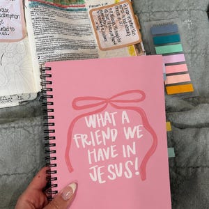 May include: A pink spiral-bound notebook with the text "WHAT A FRIEND WE HAVE IN JESUS!" in white. The cover features a pink bow design. The notebook is open to a page with text and colorful sticky notes.