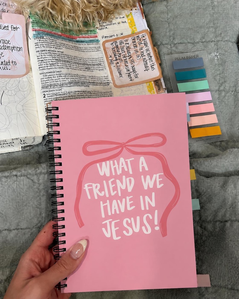 May include: A pink spiral-bound notebook with the text "WHAT A FRIEND WE HAVE IN JESUS!" in white. The cover features a pink bow design. The notebook is open to a page with text and colorful sticky notes.