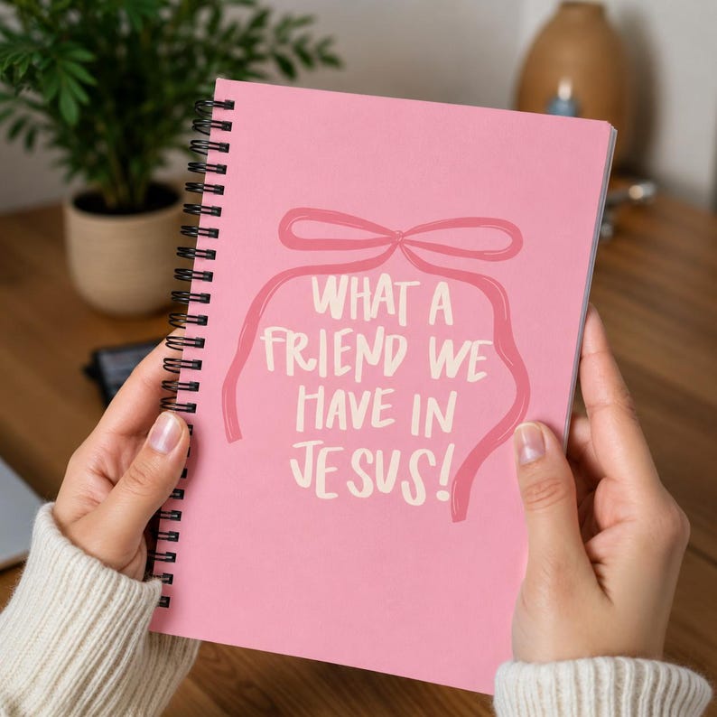 May include: A pink spiral-bound notebook with the text "WHAT A FRIEND WE HAVE IN JESUS!" in white. The text is framed by a pink ribbon design. The notebook is held open, displaying the cover.
