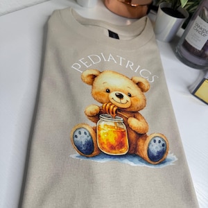 Peut inclure: T-shirt beige avec le mot "Pédiatrie" imprimé au-dessus d'une illustration à l'aquarelle d'un ours en peluche tenant un pot de miel. L'ours est brun avec des coussinets bleus, assis sur un lavis bleu.