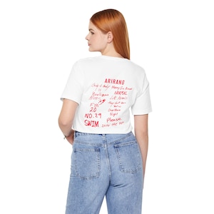 May include: White crop top with red handwritten text including "ARIRANG", "SWIM", and "NO.29". The shirt is paired with light blue denim jeans. The model is facing away from the camera.