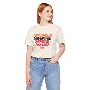 May include: Cream-colored t-shirt with the words "KEROSENE DOPAMINE CHEMICAL INDUCED" in orange, black, and red. The shirt also features a small graphic of three horizontal lines. The model is wearing blue jeans.