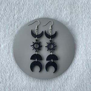 Puede incluir: Pendientes negros de temática celestial con lunas crecientes, soles y medias lunas. Los pendientes cuelgan de ganchos plateados y se exhiben sobre una superficie circular gris. Los pendientes son de un negro mate oscuro.