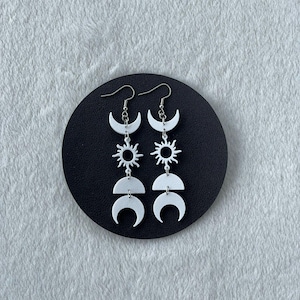 Puede incluir: Pendientes blancos de temática celestial con diseños de sol, luna creciente y media luna. Los pendientes cuelgan de ganchos plateados y se exhiben sobre una superficie circular negra.