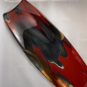 Könnte beinhalten: Rechteckige Servierplatte aus Keramik mit glänzendem Finish. Die Platte hat ein auffälliges abstraktes Design mit einer Mischung aus roten, schwarzen und blauen Farbtönen. Die Kanten sind leicht gebogen. Ein dekoratives Stück.