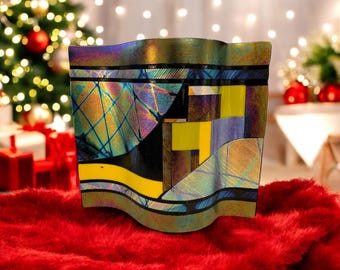 May include: A decorative glass vase with a wavy top edge. The vase features a geometric design with yellow, blue, and black shapes. The glass has iridescent qualities, reflecting multiple colors. The vase sits on a red, fuzzy surface.