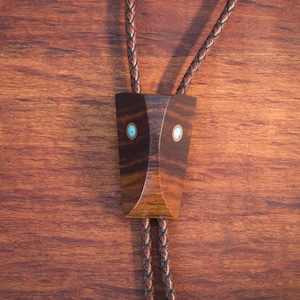May include: A bolo tie with a geometric wooden pendant. The pendant is dark brown with turquoise and white inlays. The braided leather cord is a matching brown color. The tie is displayed on a wooden surface.
