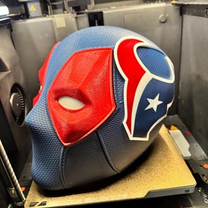 Könnte beinhalten: Ein 3D-gedruckter Helm mit blauem und rotem Design. Der Helm hat einen Stern und ein rot-weißes Logo an der Seite. Der Helm steht auf einer braunen Plattform in einem 3D-Drucker.