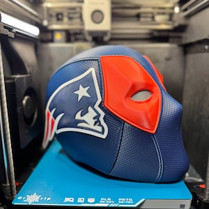 Könnte beinhalten: Ein blau-roter Helm mit einem weißen Stern und Logo wird auf einer blauen Oberfläche in einem 3D-Drucker angezeigt. Der Helm hat eine strukturierte Oberfläche und eine rote Augenmaske. Das Logo ist weiß und blau.