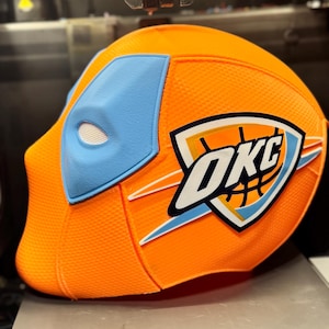 Op de afbeelding: Een oranje en lichtblauw masker met het OKC-logo, waarschijnlijk met een sportthema. Het masker heeft een textuur en een witte oogopening. Het OKC-logo toont een basketbal en de letters "OKC".