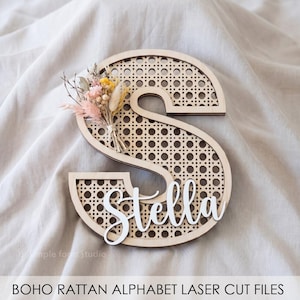 Puede incluir: Una letra "S" de madera con diseño de ratán y el nombre "Stella" en escritura blanca. La letra está decorada con un pequeño ramo de flores secas en rosa, amarillo y beige. El texto "BOHO RATTAN ALPHABET LASER CUT FILES" está en la parte inferior.