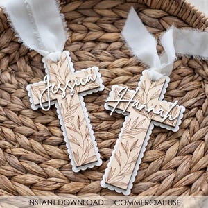 May include: Two wooden cross decorations, each with a name in white script: "Jessica" and "Hannah". The crosses feature a leaf design and scalloped edges, with white ribbons. The background is a woven basket.