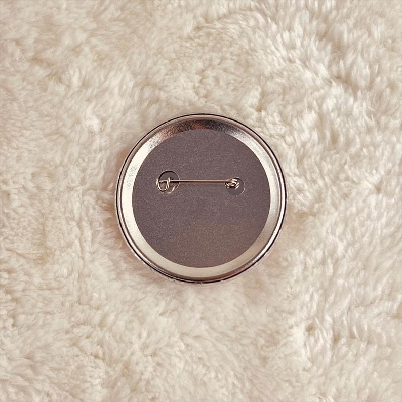 May include: The back of a round button with a silver pin and a gray backing.