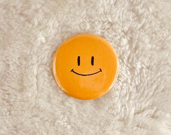 May include: A yellow smiley face button with a black outline. The button is on a white fuzzy surface.