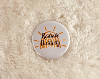 May include: A white button with a rainbow and sun design. The text "Radiate Positivity" is written in black.