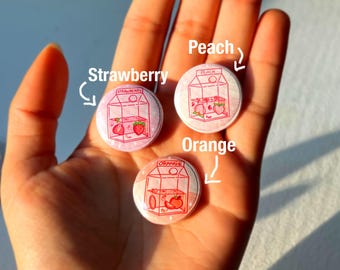 May include: Three round pins with a cartoon illustration of a milk carton with different fruits inside. The pins are labeled "Strawberry", "Peach", and "Orange".