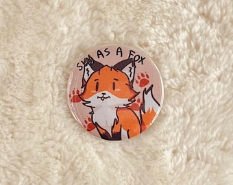 May include: A round button with a pink background and a cartoon fox with orange fur and black eyes. The fox is smiling and has a white chest. The text "Sly as a Fox" is written in black around the fox.